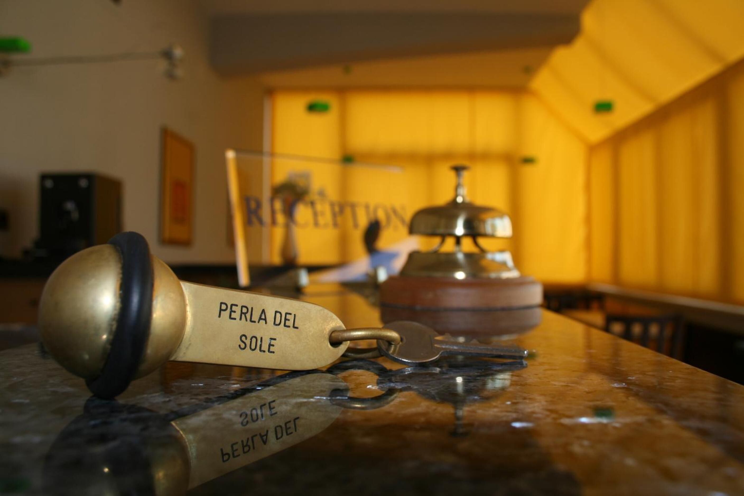 A brass key with "PERLA DEL SOLE" engraved on it, lying on a marble surface in a warmly lit room with wooden paneling.