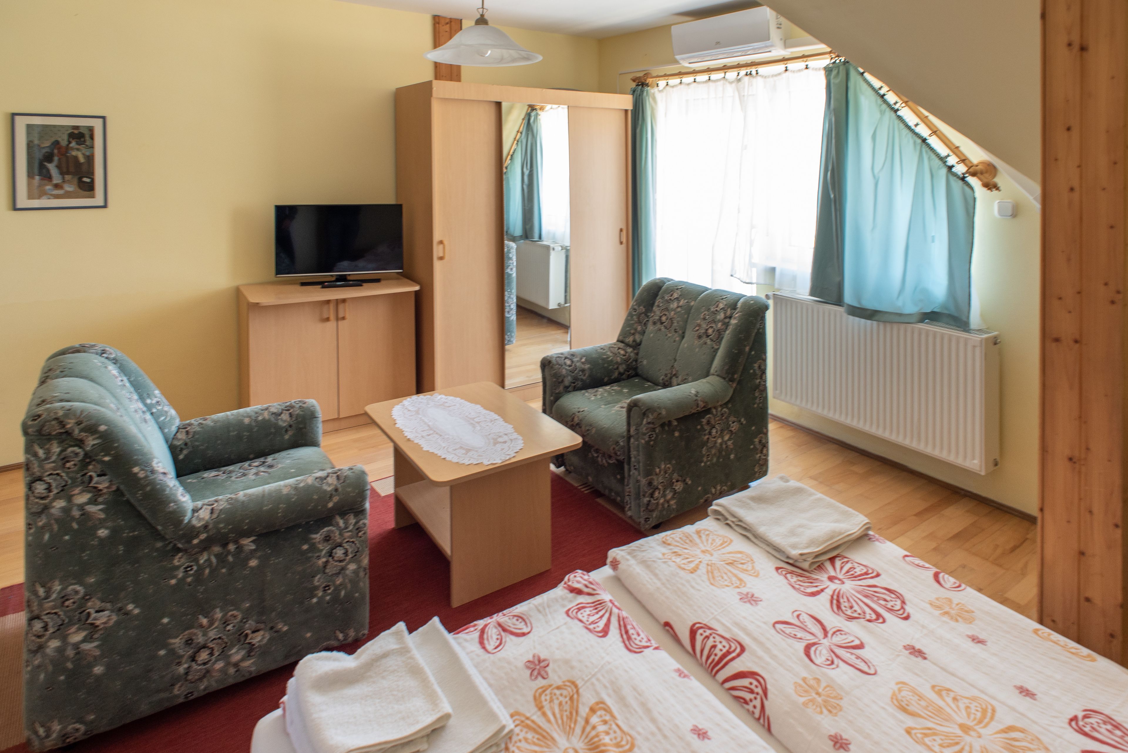 Een kamer met twee fauteuils met bloemenprint, een klein houten tafeltje met een witte tafelkleed, een bed met een bloemenbedekker, een televisie op een houten kast, en een raam met gordijnen.