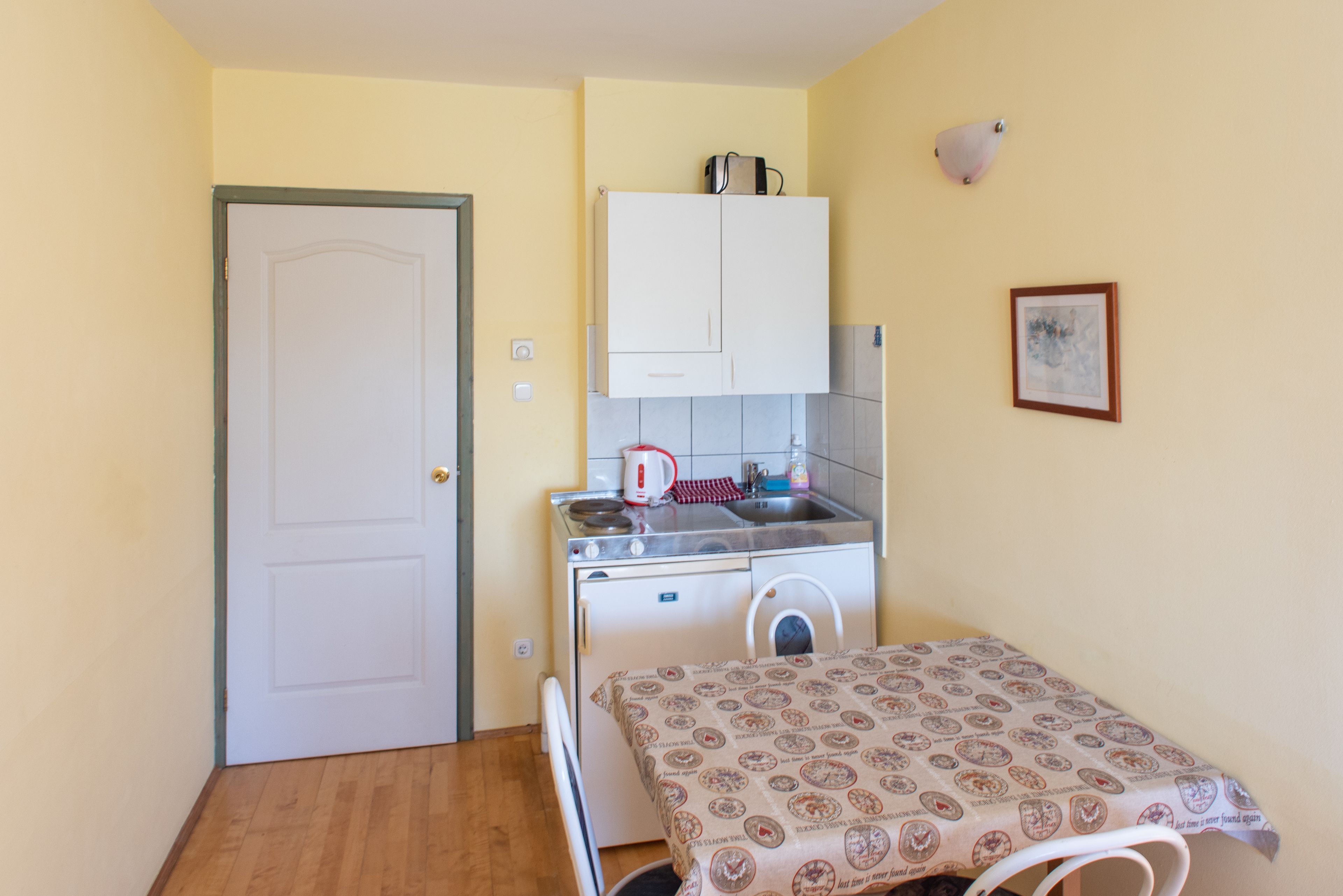Una cocina compacta con un refrigerador blanco, un lavamanos y un microondas. Hay una mesita con mantel estampado y una silla.