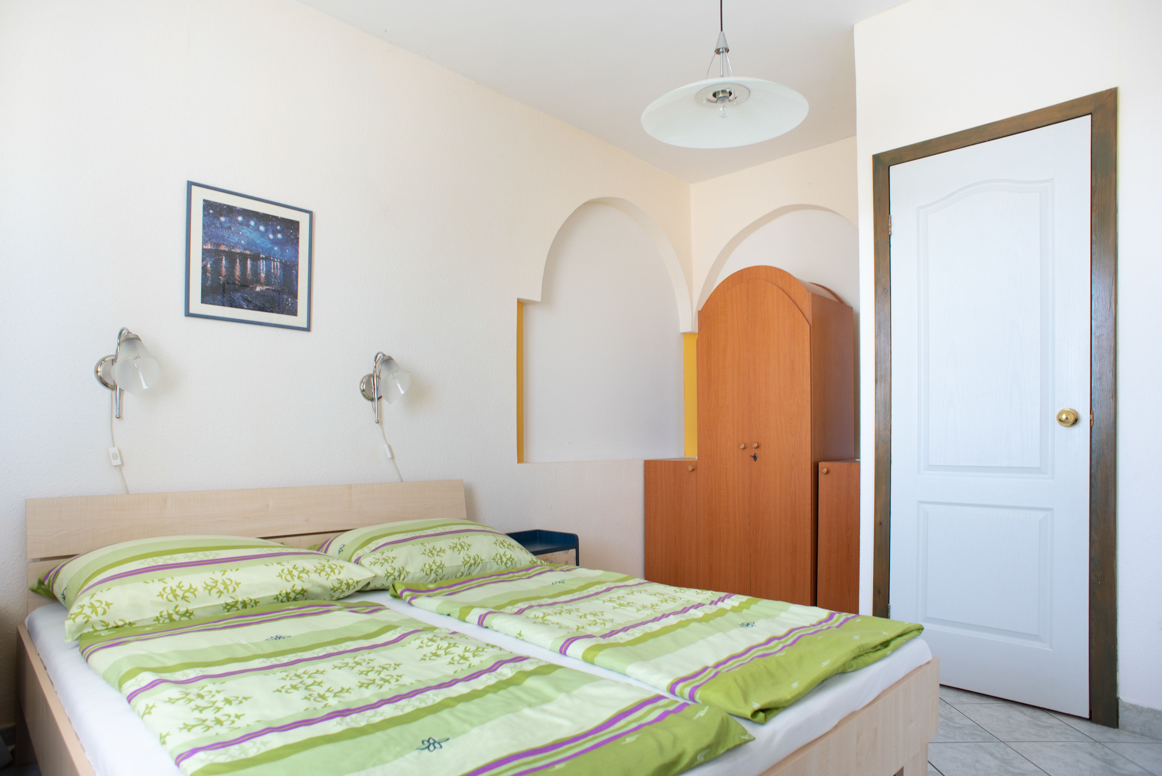 A bedroom with a bed, a nightstand, a wooden wardrobe, a door, and a wall-mounted picture.