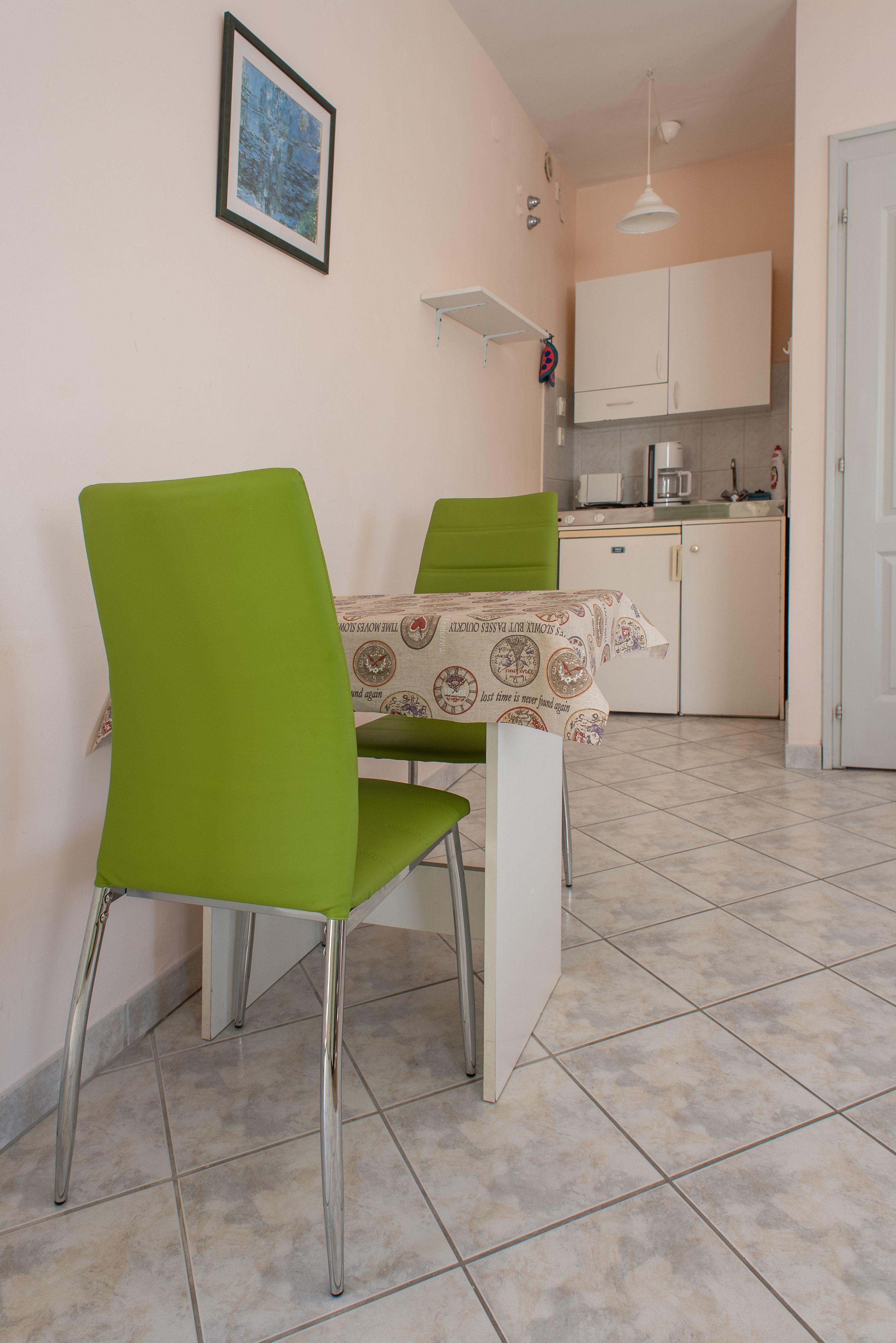 Una cocina pequeña con armarios blancos, un microondas y una cafetera. Hay dos sillas verdes y una mesa con mantel estampado.