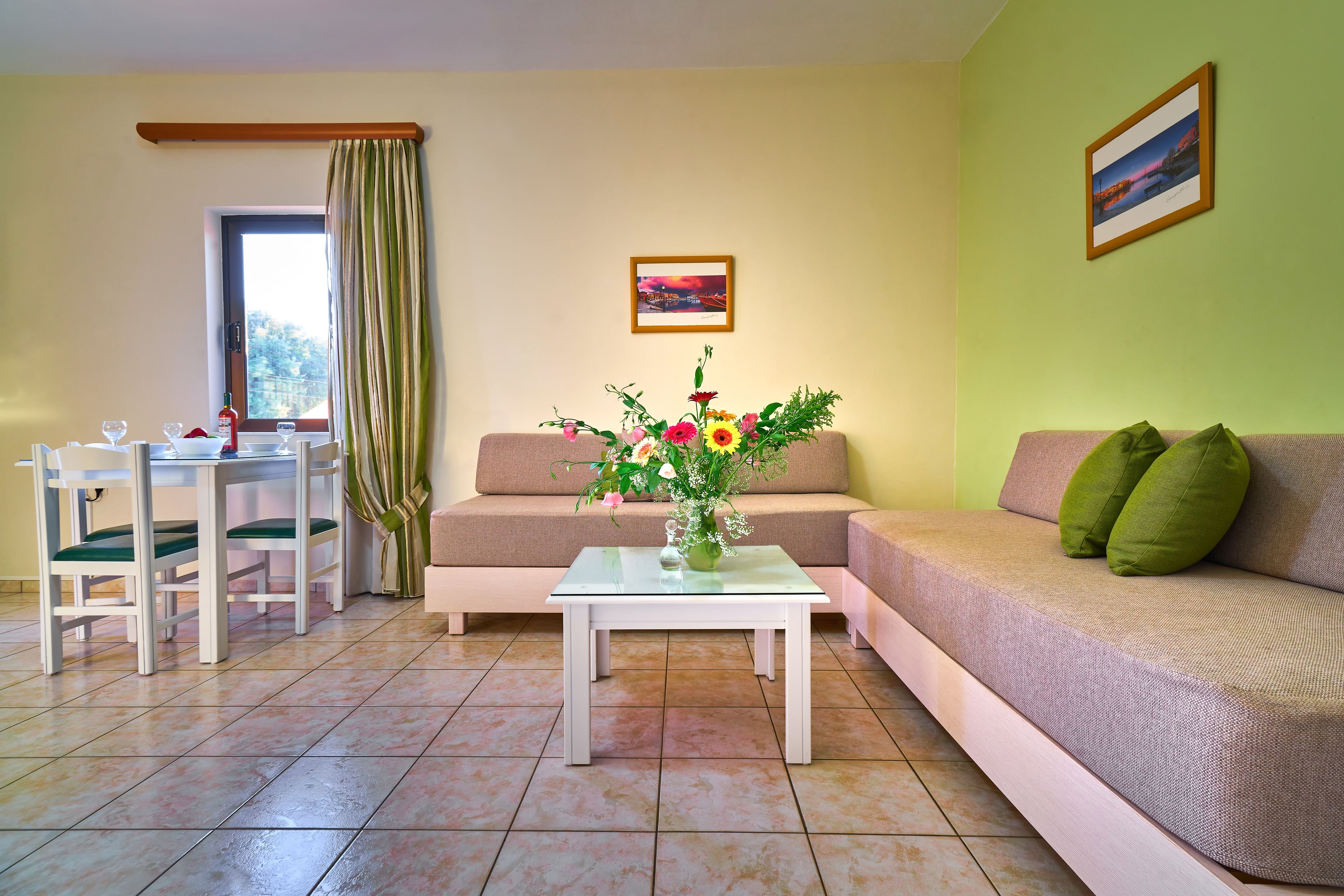 Een woonkamer met een bank, een glazen salontafel met bloemen, een eettafel met stoelen en een raam met gordijnen.