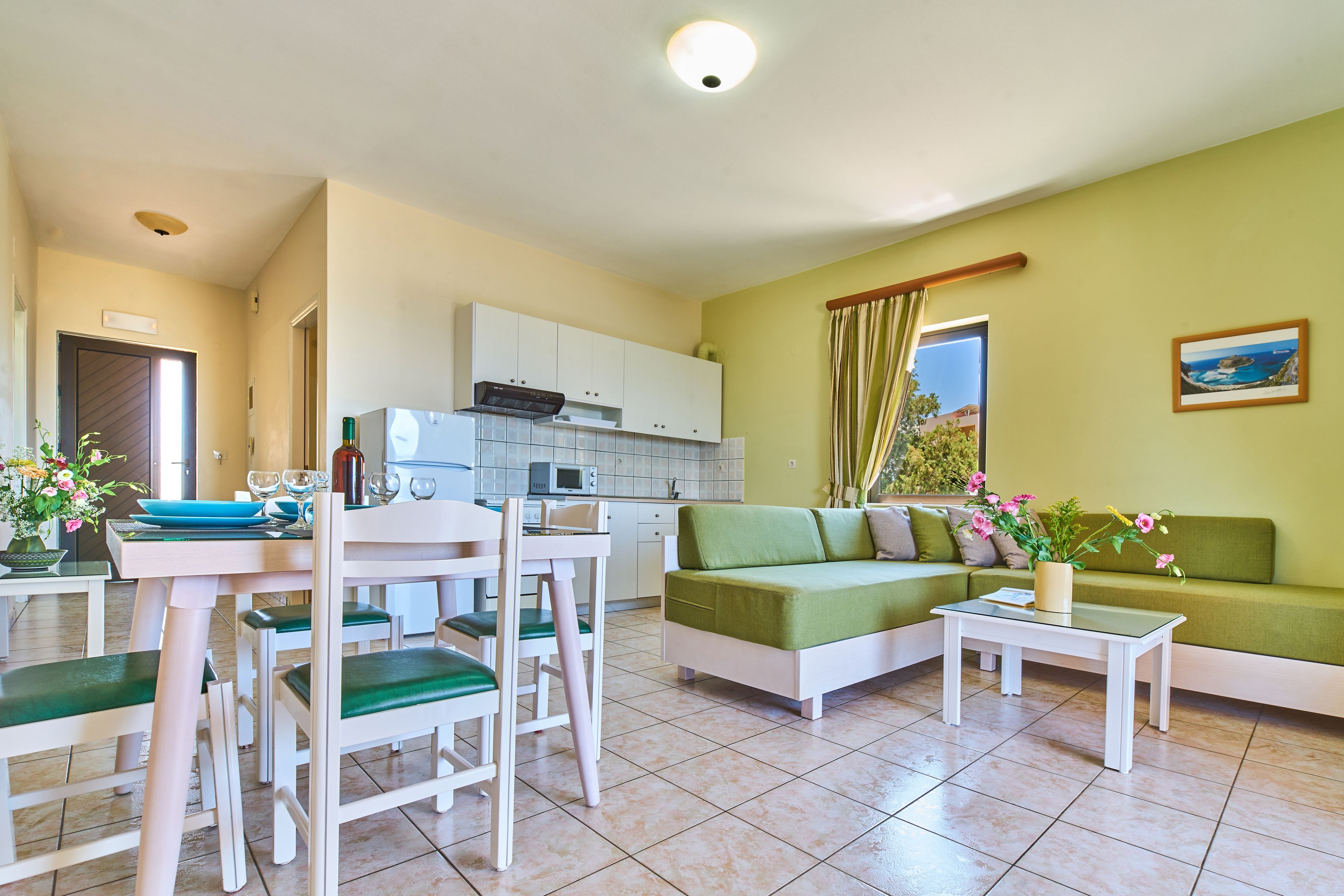 Una sala de estar moderna con un sofá verde, una mesa de centro blanca y sillas a juego. Se ve una cocina al fondo con armarios blancos y un microondas.