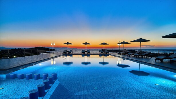 Uma área de piscina com espreguiçadeiras e guarda-sóis, piscina com azulejos e vista para o pôr do sol.