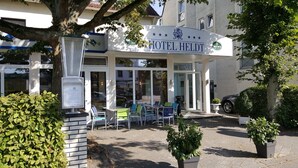 En hotelindgang med et skilt, der siger "HOTEL HELDT", udendørs siddepladser med stole og en potteplante i forgrunden.