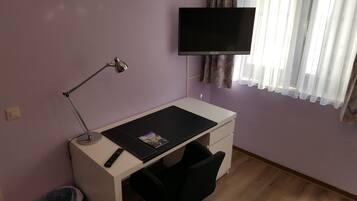 Un pequeño estudio con escritorio, silla, lámpara, televisor y ventana con cortinas.