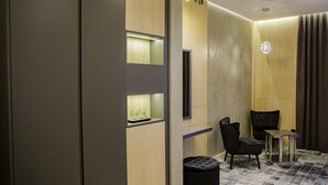 A modern hotel room with a seating area featuring a black upholstered chair, a small table, and a black ottoman. The room has a wooden accent wall, a large window, and a patterned rug on the floor. - VisPas Hotel (Chisinau)