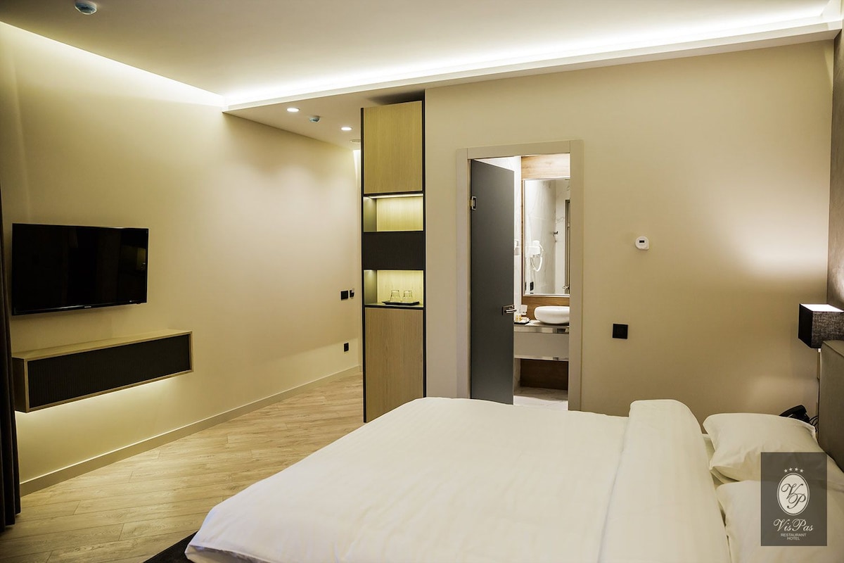 A modern hotel room with a large bed, a flat-screen TV mounted on the wall, a bedside table, and a walk-in closet.