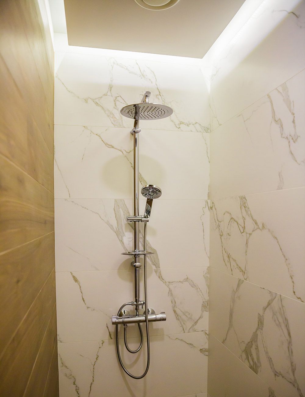A shower with a handheld showerhead and a handheld faucet, set against marble walls.
