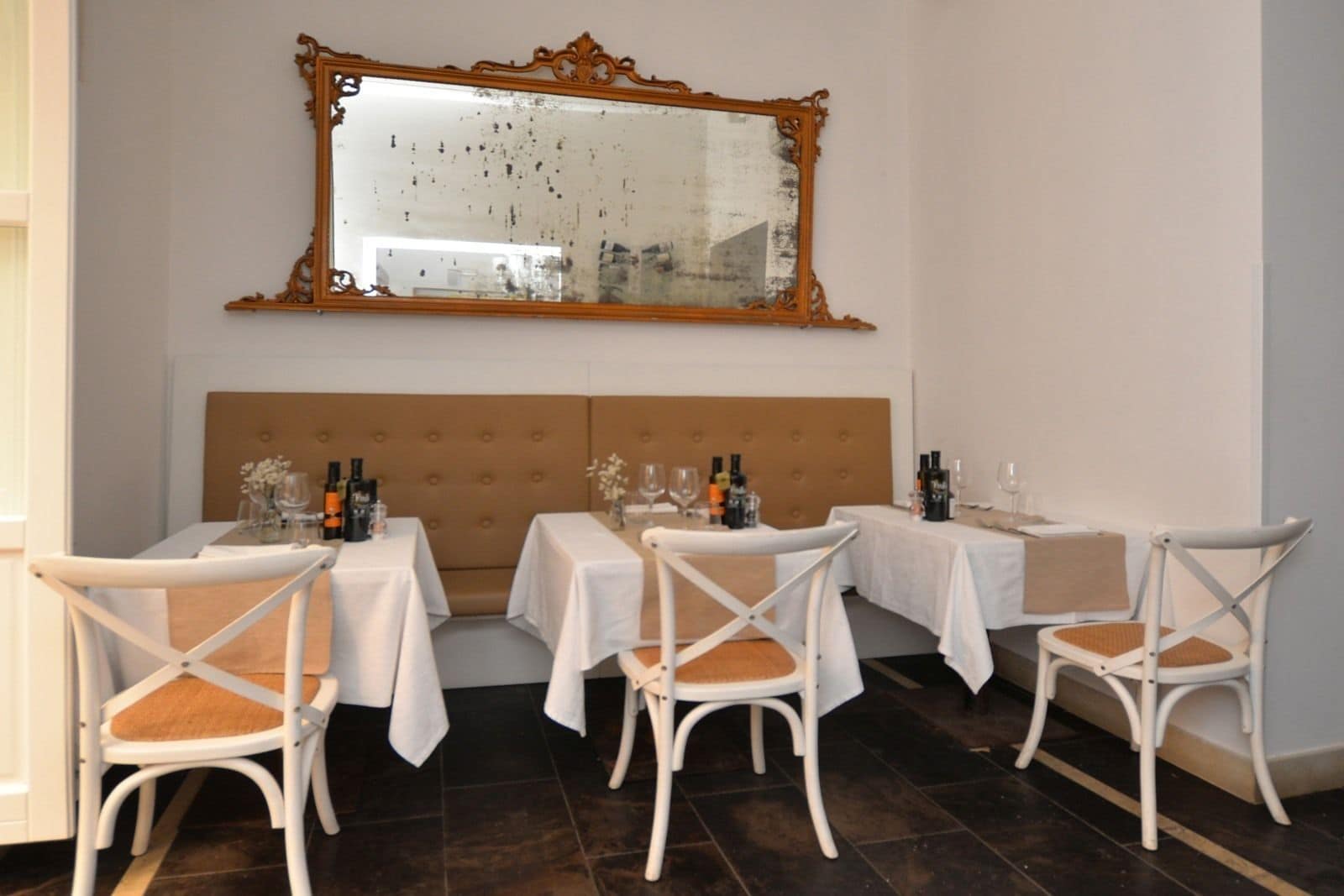 A dining area with four tables, each with white tablecloths and white chairs. There is a large mirror above the tables, and a tufted headboard behind them.