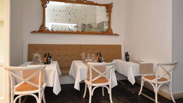 A dining area with four tables, each with white tablecloths and white chairs. There is a large mirror above the tables, and a tufted headboard behind them.