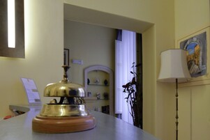 Zona de recepción de hotel con mostrador de madera, un timbre, escritorio con computadora y una lámpara.