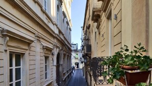 Una calle estrecha flanqueada por edificios, con una fila de mesas y sillas a la derecha, lo que sugiere un ambiente de café o restaurante.