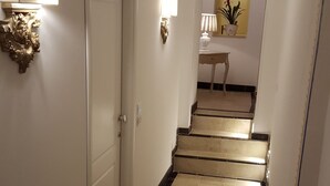 Un pasillo con una puerta, apliques en la pared, una mesita con una lámpara y una escalera que conduce a otra habitación.