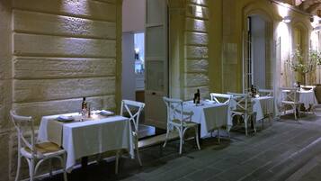 Un espace extérieur aménagé pour les repas, avec des tables et des chaises blanches, des murs en pierre et un sol pavé.