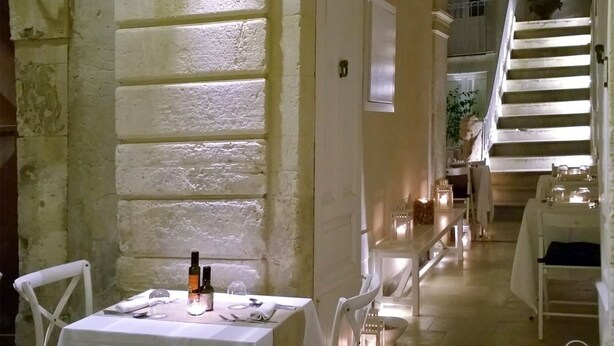 Un área de comedor al aire libre con mesas y sillas blancas, una escalera que lleva a un nivel superior y una planta en maceta en la escalera.