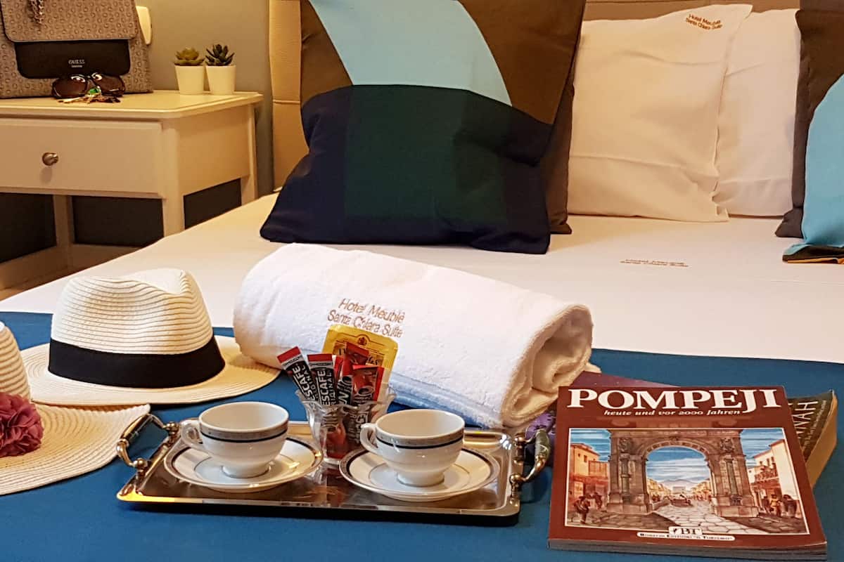 A neatly arranged hotel room with a bed, pillows, a nightstand, a tray with cups and a hat, and a book titled "POMPEJI".