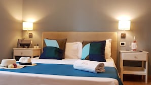 A neatly made bed with a blue bedspread, two pillows, and a folded towel. There are two bedside tables, one with a lamp and the other with a bottle and glasses. - Hotel Meublè Santa Chiara Suite (Naples)