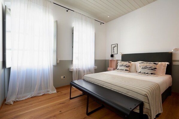 Une chambre à coucher comprenant un lit, un banc, des tables de chevet, des rideaux et un luminaire fixé au mur.
