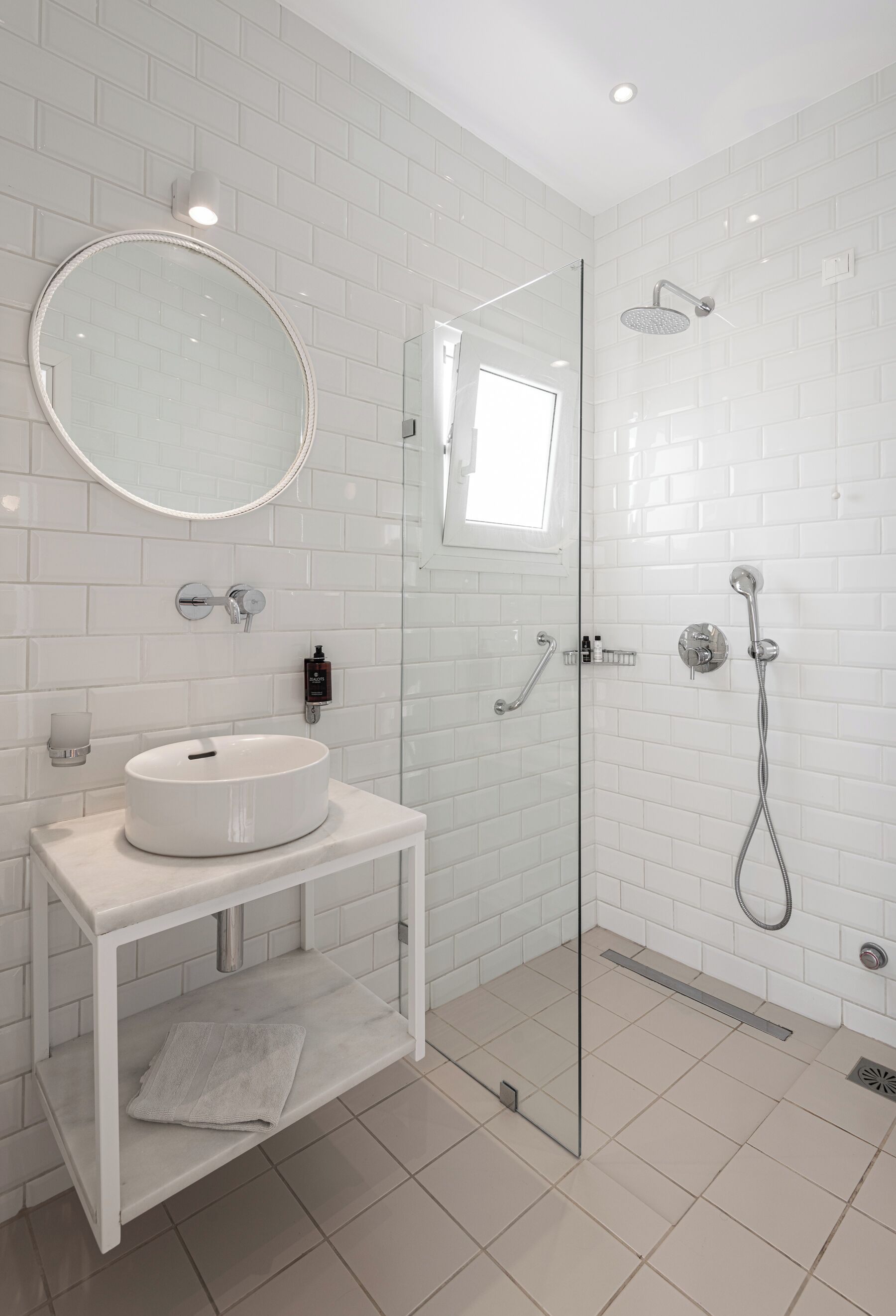 A modern bathroom with a white tiled wall, a round mirror, a white sink, and a glass-enclosed shower area.