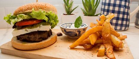 A burger with fries on a wooden board, a bowl of sauce, and two potted plants.