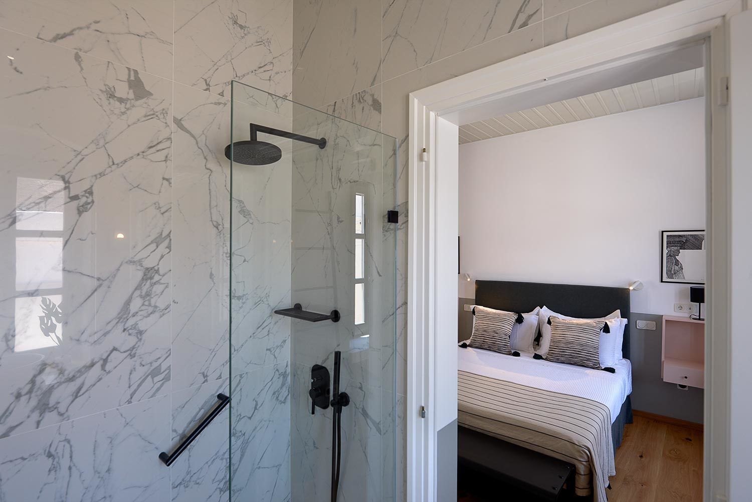 A modern bathroom with a glass-enclosed shower, marble walls, and a visible bed with striped bedding and pillows.