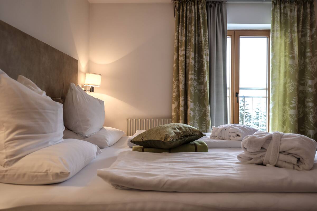A hotel room with a neatly made bed, a bedside lamp, a window with curtains, and a radiator.