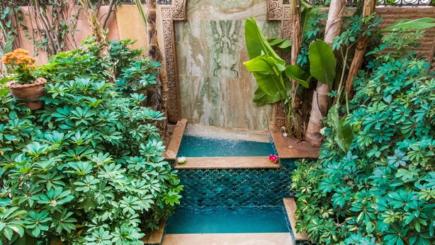 A stone staircase leading to a pool surrounded by lush greenery and a decorative archway. - Riad Mazal (Marrakech)