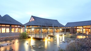 A resort with thatched-roof buildings, a wooden bridge over a calm water body, and a stone wall. - Garden Route Game Lodge (Albertinia)