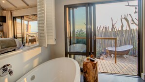 A modern bathroom with a large bathtub, a wooden stool, and a view of a wooden deck and outdoor furniture. - Garden Route Game Lodge (Albertinia)