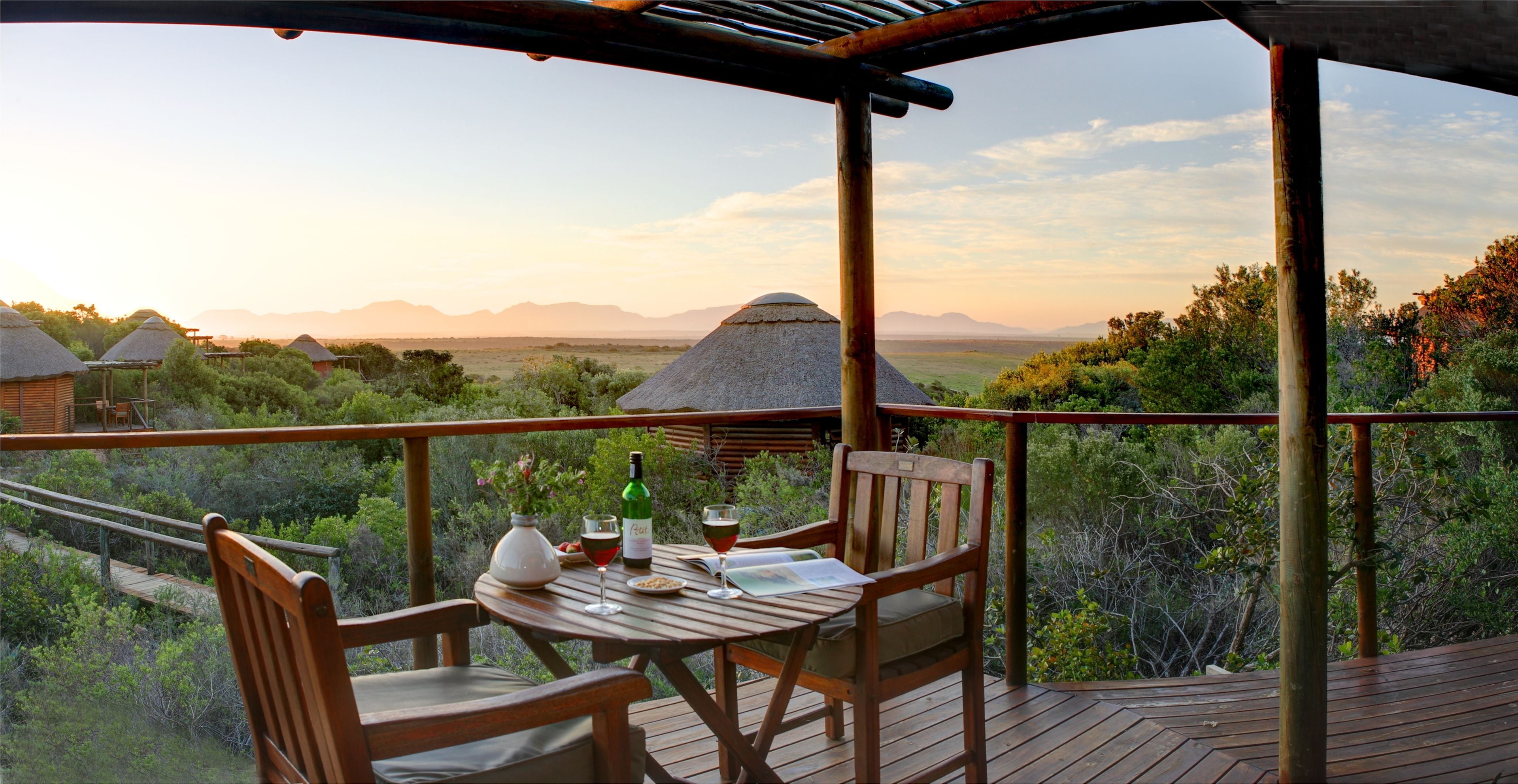 A wooden deck with a table set for two, overlooking a savanna landscape with thatched-roof huts and distant mountains.