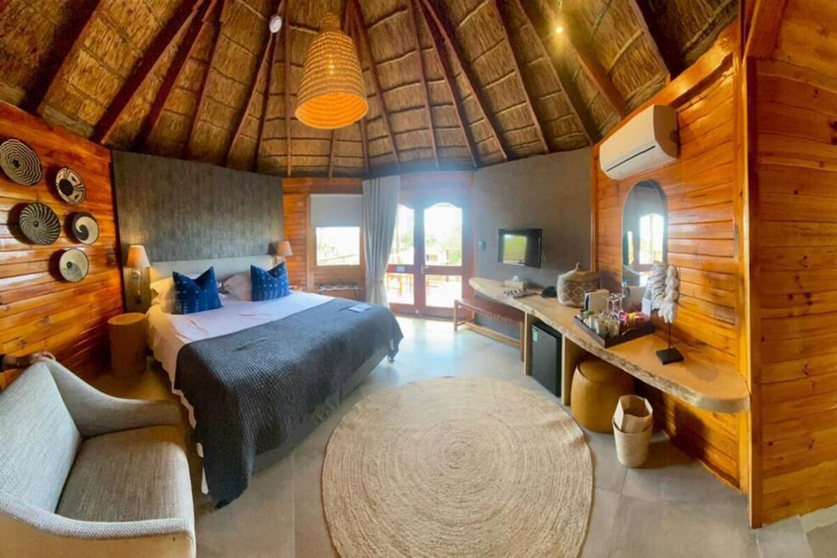 A wooden cabin with a thatched roof bedroom, a sitting area with a sofa and a round rug, and a bathroom with a sink and mirror.