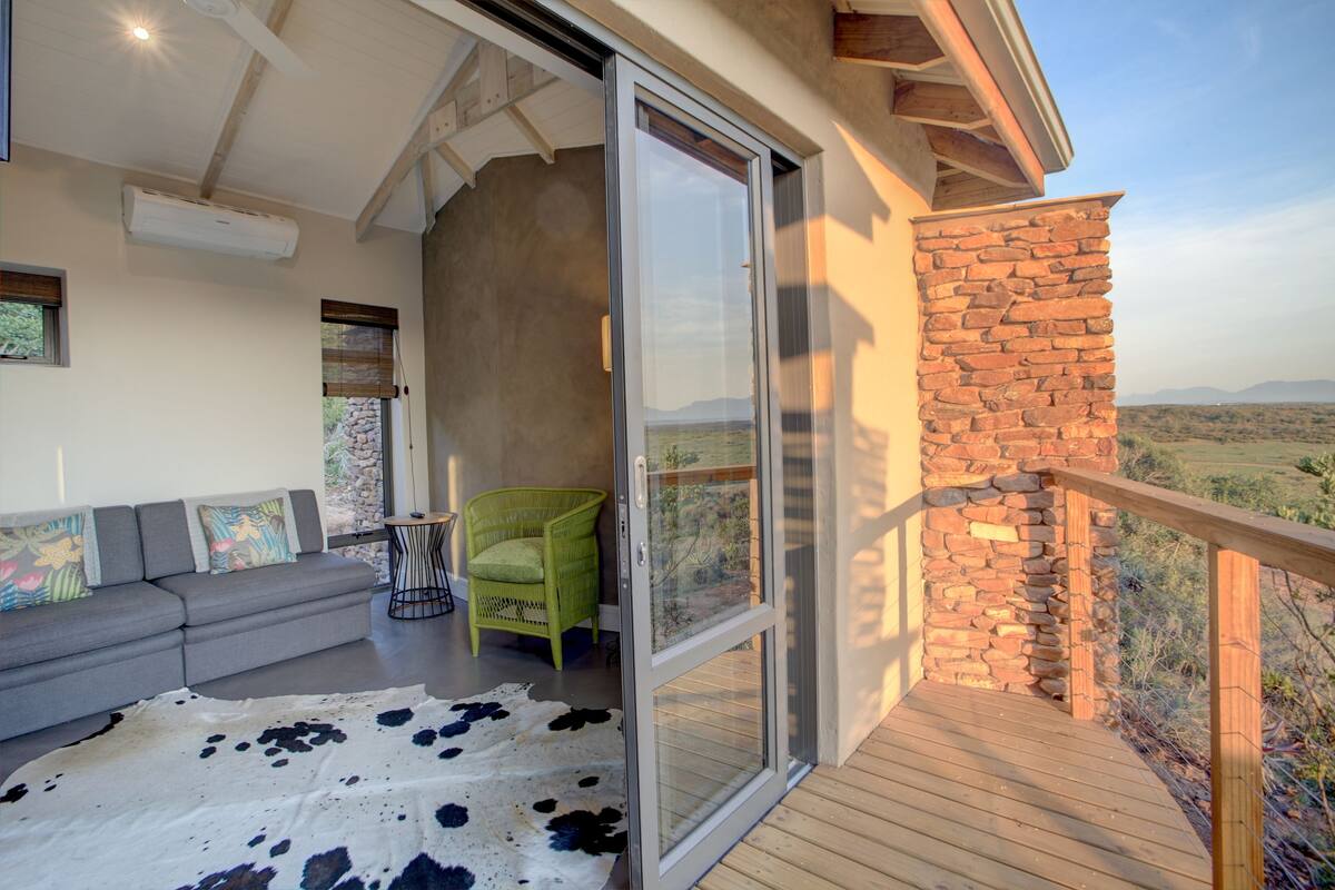 A balcony with a stone wall, a wooden deck, a grey sofa, a green chair, and a cowhide rug.