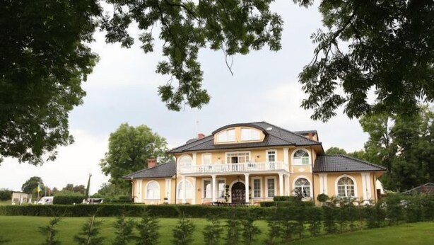 Een groot, twee verdiepingen tellend huis met een balkon, omringd door een verzorgd gazon en bomen.