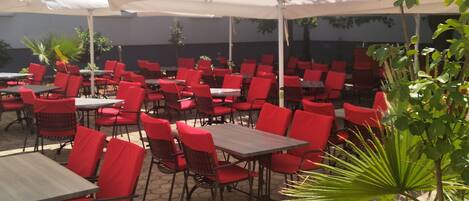 An outdoor seating area with red chairs and white umbrellas, arranged on a paved patio.