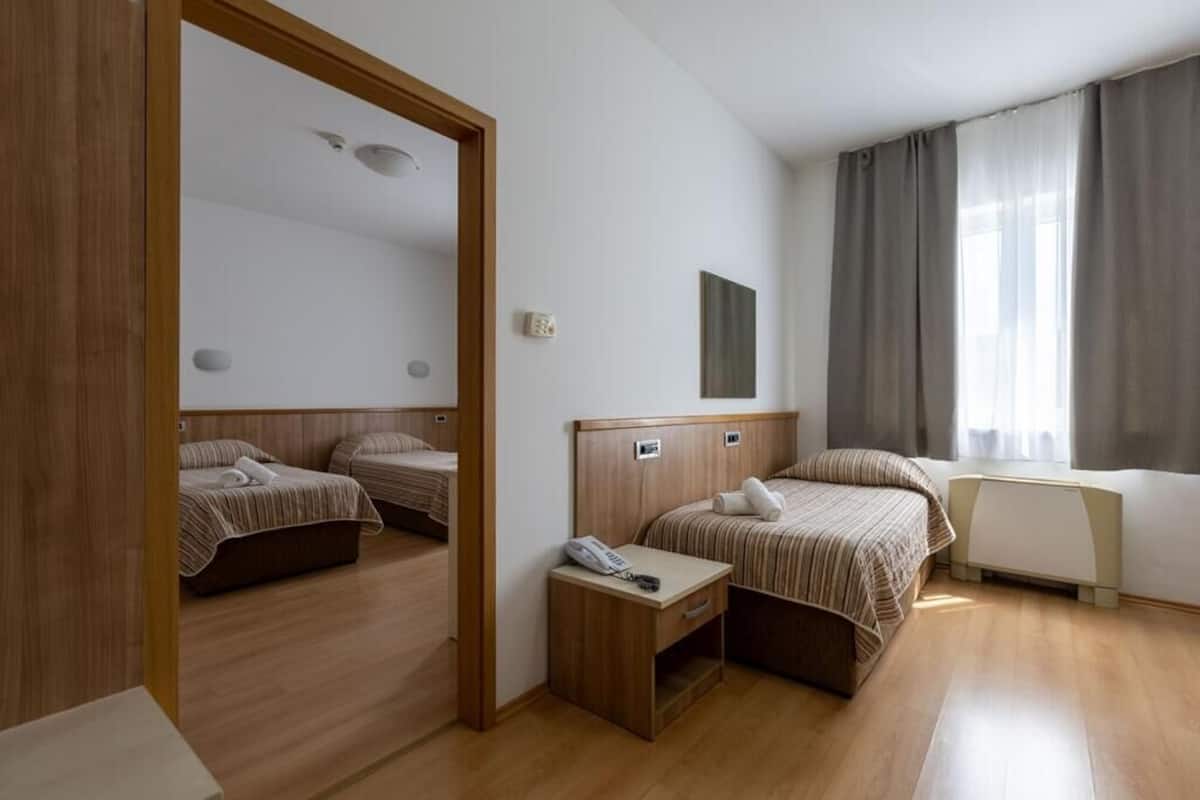 Une chambre d’hôtel avec deux lits, un plancher en bois, une table de chevet avec un téléphone, un radiateur et une fenêtre avec des rideaux.