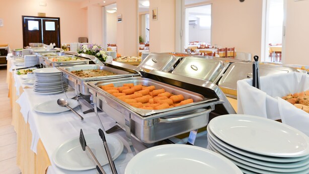 A buffet setup with multiple food stations, including trays of tater tots and other dishes, arranged on a long table with stacked plates and serving utensils. - Hotel Porto (Zadar)