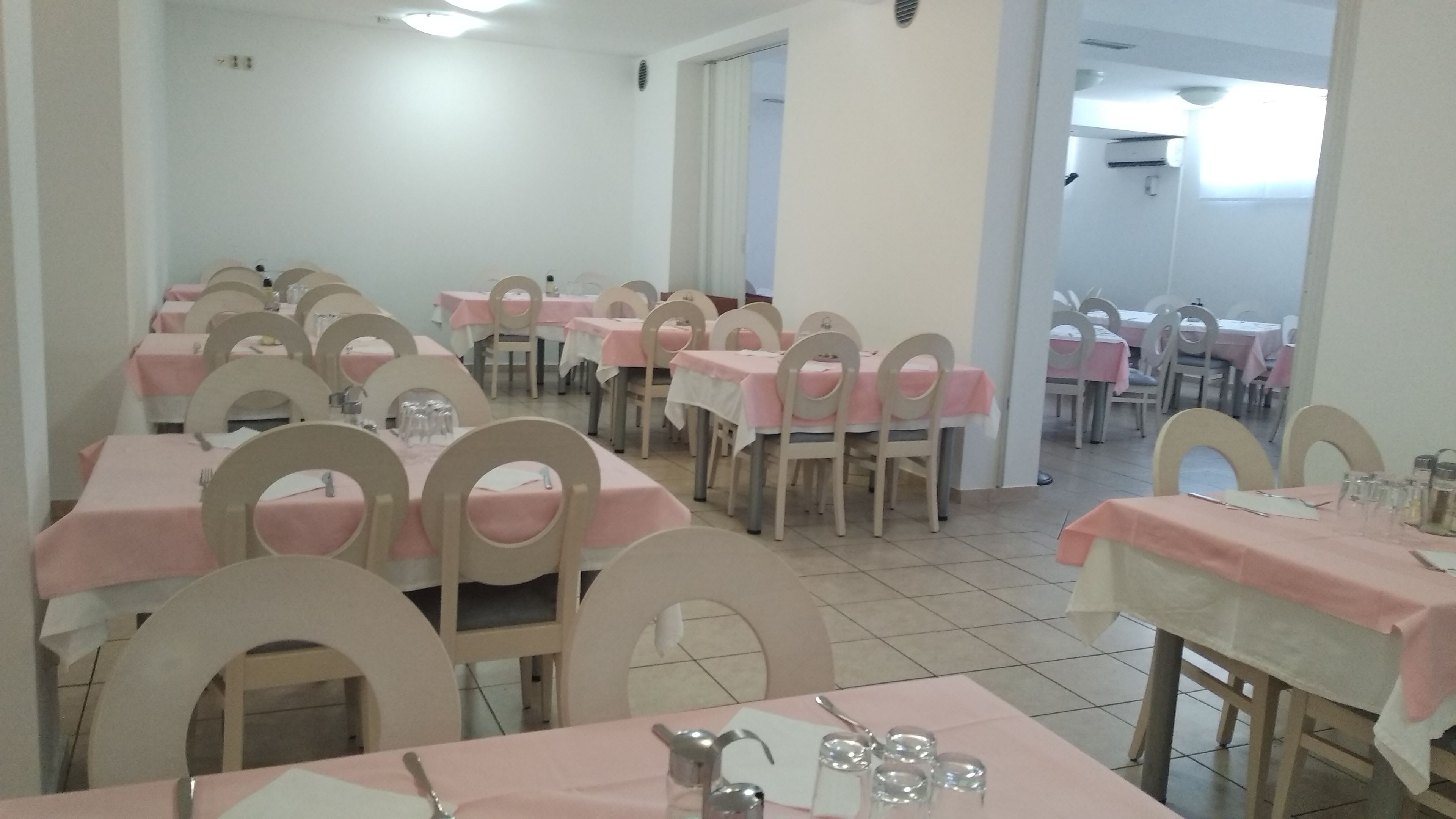 Ein Essbereich mit Tischen und Stühlen, einige Tische sind mit rosa Tischdecken bedeckt, und der Raum hat weiße Wände sowie Fliesenboden.