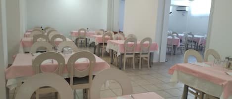 A dining area with tables and chairs, some tables have pink tablecloths, and the room has white walls and tiled flooring.