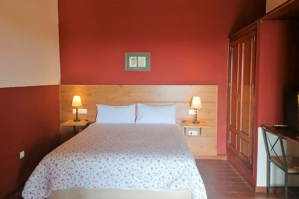 Ein Hotelzimmer mit hölzernem Kopfteil, zwei Nachttischlampen, einem Bett mit floralem Bettbezug, einem Nachttisch aus Holz und einem Kleiderschrank.