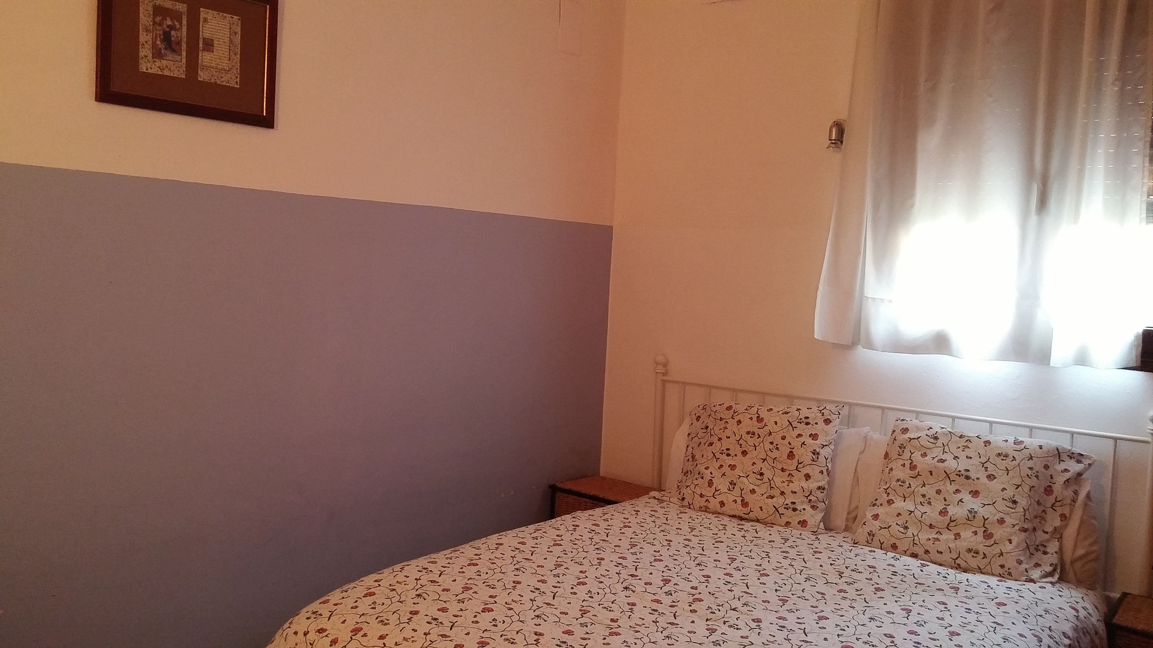 A bedroom with a bed, a nightstand, a window with curtains, and a wall-mounted picture frame.