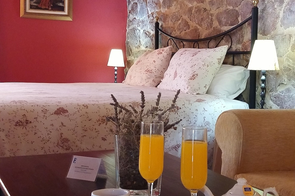 A hotel room with a stone wall, a bed with floral bedding, a red wall with a framed picture, and a breakfast tray with croissants and orange juice.