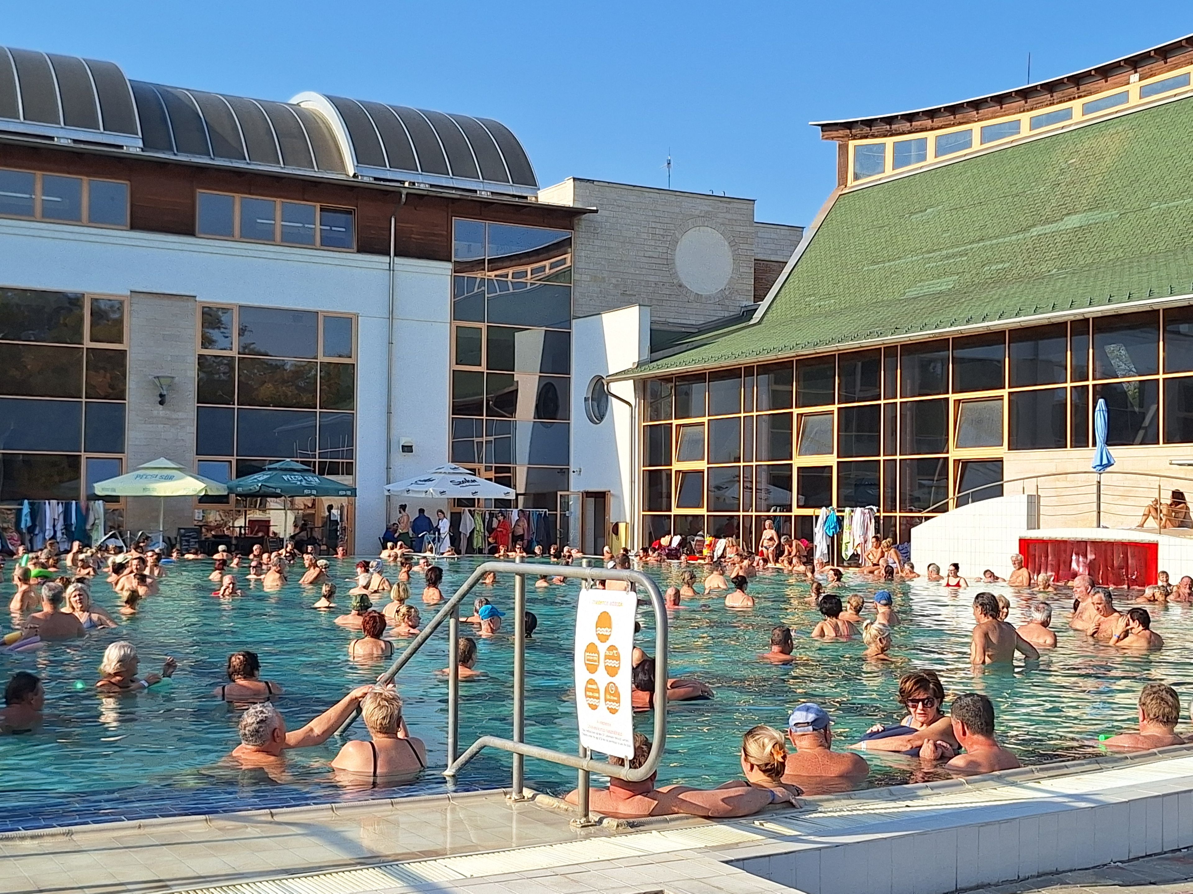 Zona de piscina con varias personas, un puesto de socorrista y un edificio con grandes ventanales.