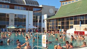 Zona de piscina con varias personas, un puesto de socorrista y un edificio con grandes ventanales.