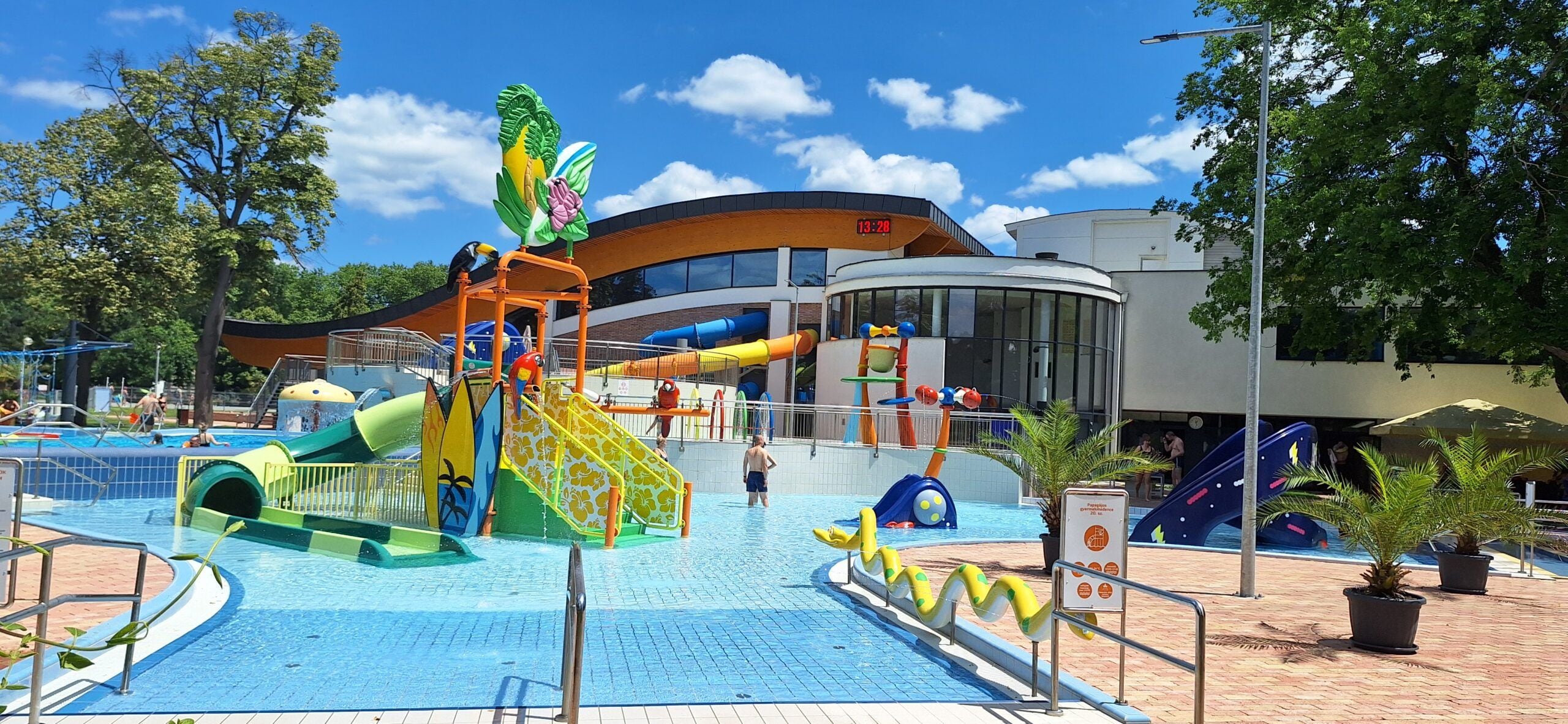Parque acuático con toboganes y estructuras de juego, rodeado de árboles y un edificio con un reloj.
