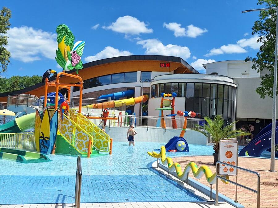 Parque acuático con toboganes y estructuras de juego, rodeado de árboles y un edificio con un reloj.