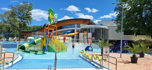 Parque acuático con toboganes y estructuras de juego, rodeado de árboles y un edificio con un reloj.