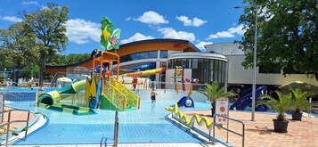Parque acuático con toboganes y estructuras de juego, rodeado de árboles y un edificio con un reloj.