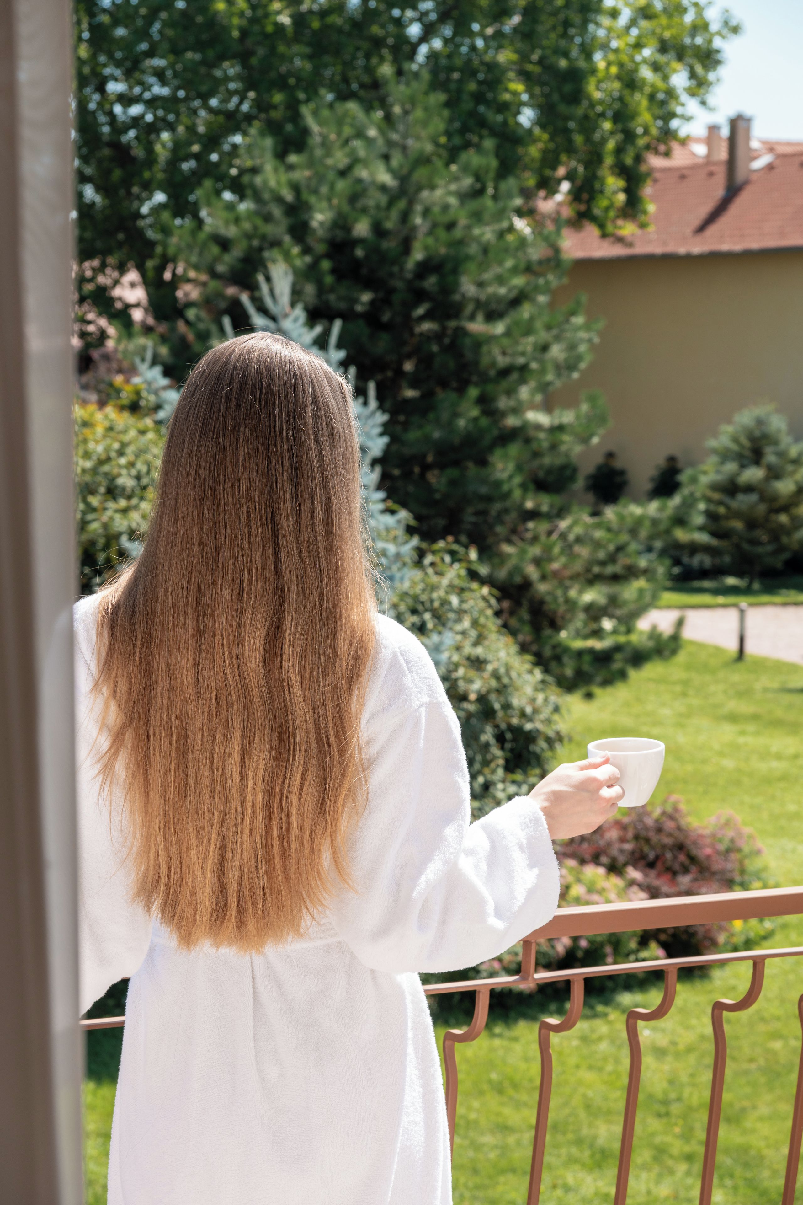 Una persona de cabello largo parada en un balcón sosteniendo una taza, con un jardín y un edificio al fondo.
