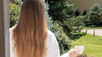 Una persona de cabello largo parada en un balcón sosteniendo una taza, con un jardín y un edificio al fondo.